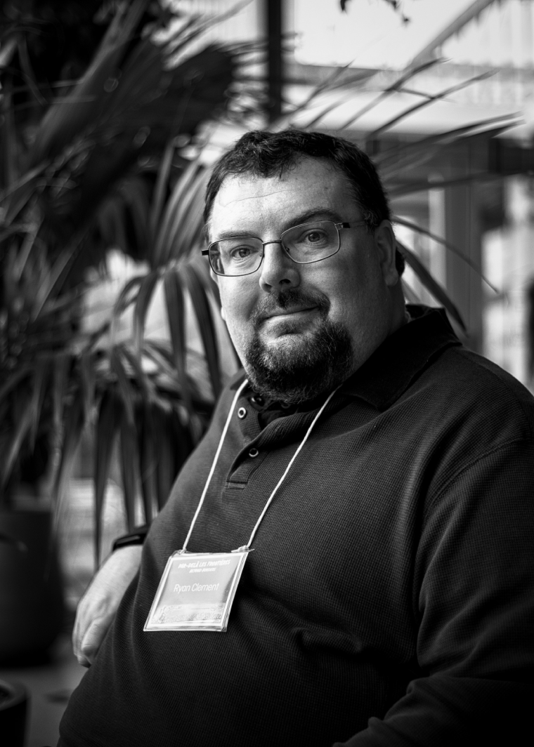 A black and white photo of a person looking at a camera. Large plant in background.