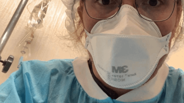 Person in a hairnet, mask and glasses in a blue medical gown looking down at camera. IV pole in the background.