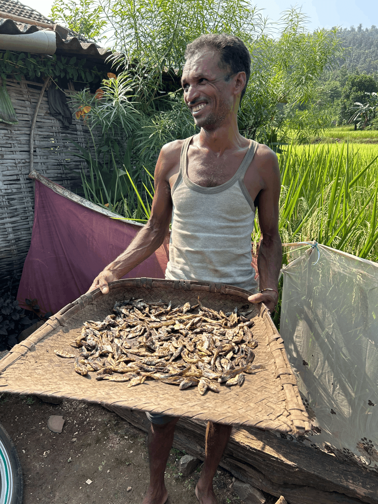 A person smiling, standing outdoors holding a woven tray filled with dried fish.