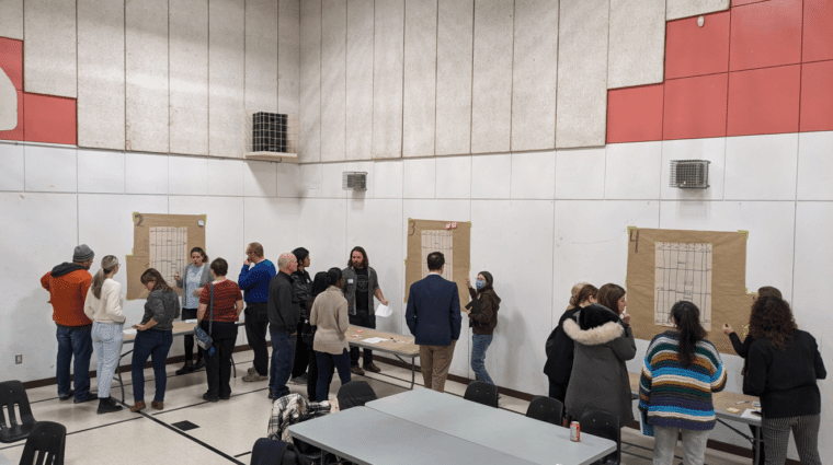 People standing around tables looking at displays and discussing in a gymnasium.