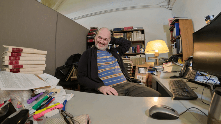 Person at a desk in an office with books and bookcases, papers, highlighters, pens and a pile of books on desk.