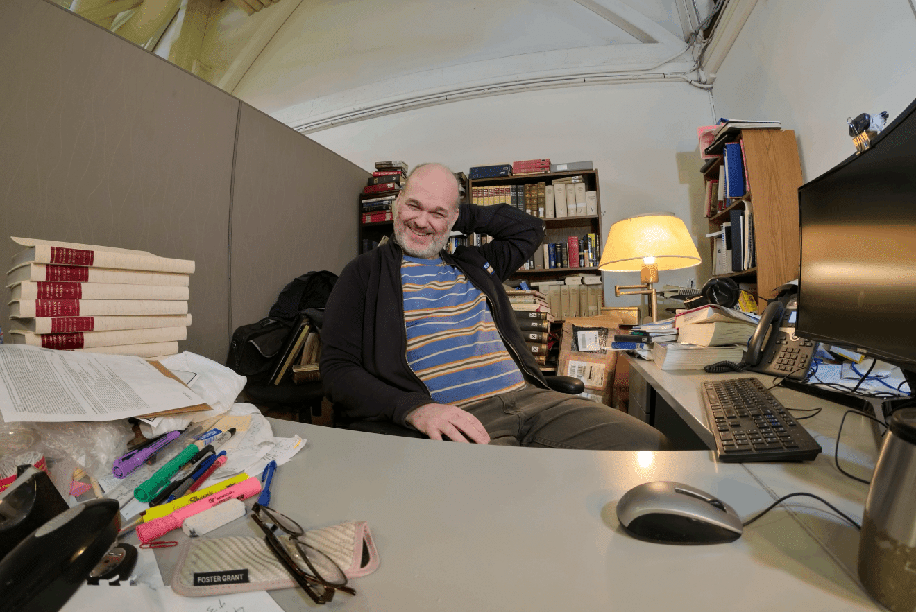 Person at a desk in an office with books and bookcases, papers, highlighters, pens and a pile of books on desk.