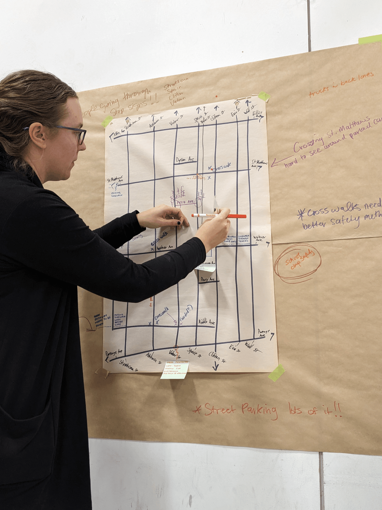 A person stands at a board looking at a paper with a grid like drawing. The person is holding a red marker.