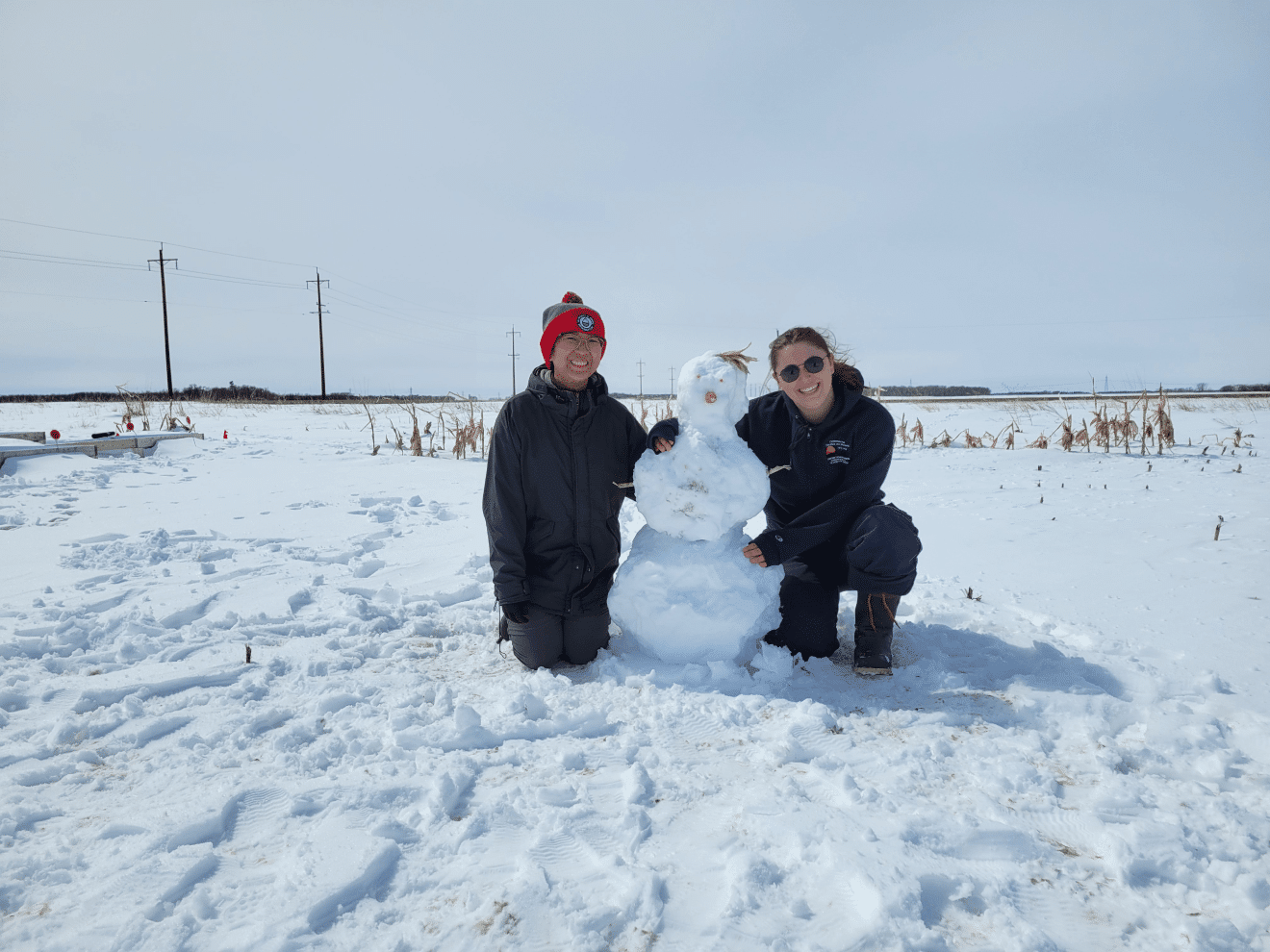 Two people in a field sitting next to small snowman.
