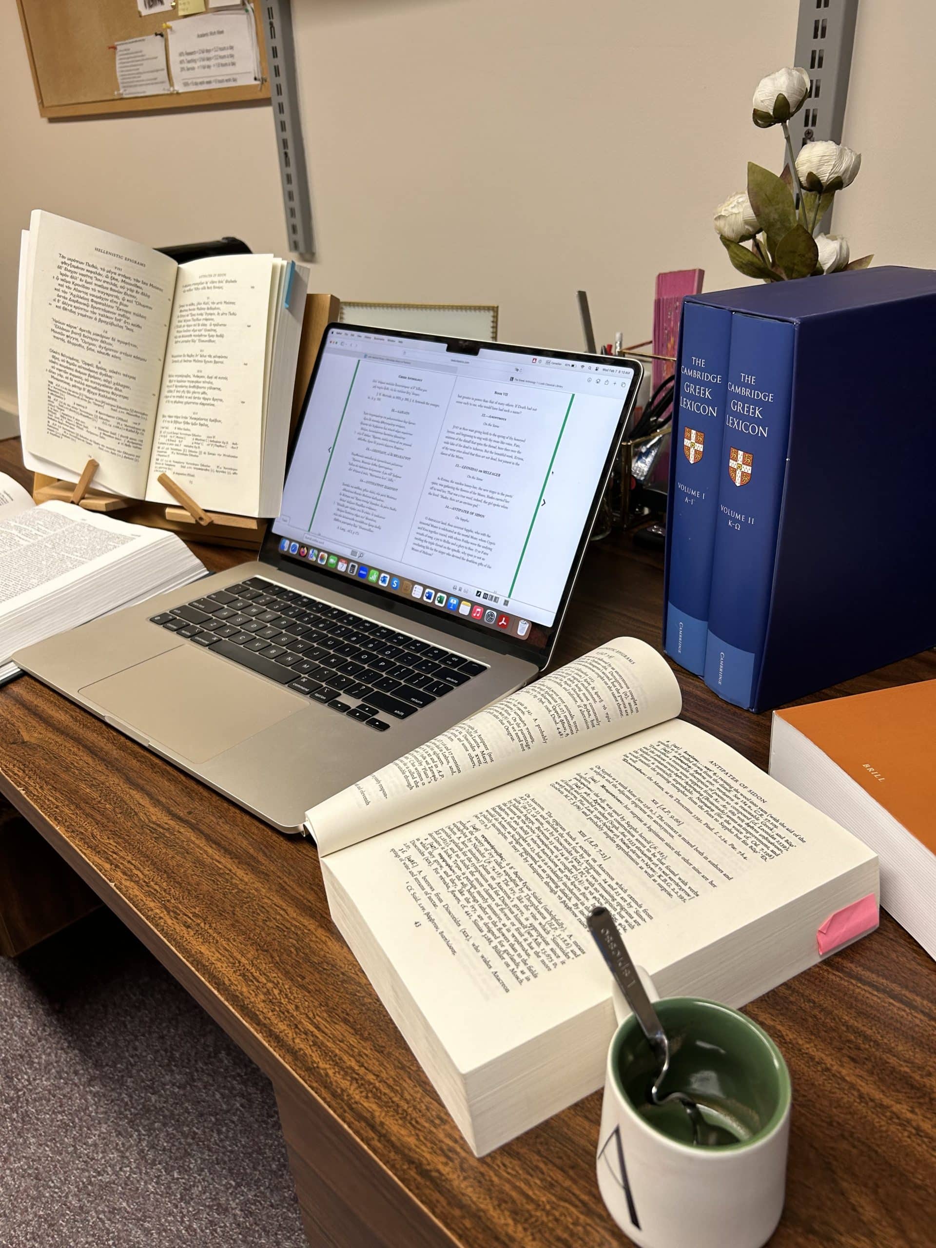 A laptop at a desk, surrounded by three open books and a cup.