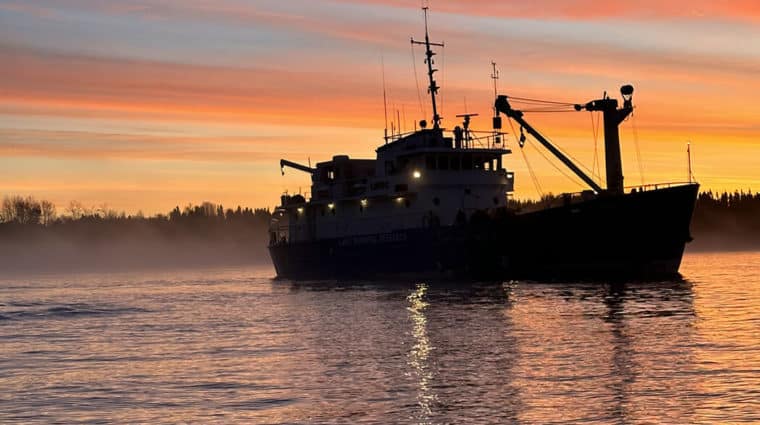 A coloured photo of a large ship with light contrast making in appear shadowed In the background a beautiful sunrise with mist hovering over the water