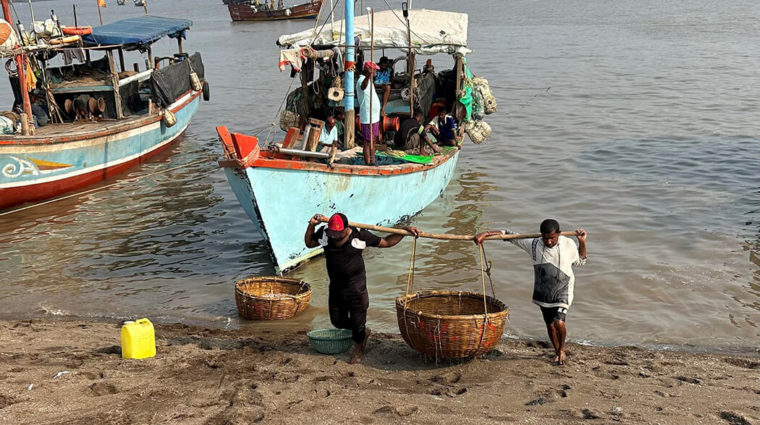 Two people carrying a big bamboo basket together with boats in the background