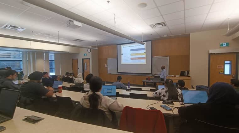 University classroom with students attending a lecture on relational databases. The professor presents a slide while students take notes on laptops.
