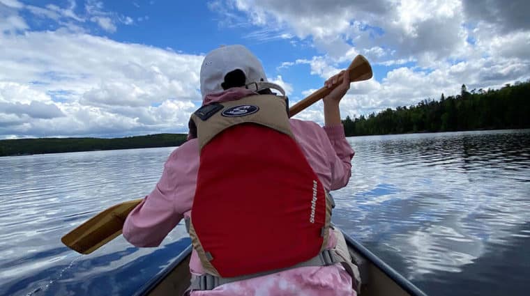 A person sitting in a canoe, canoeing across a lake, with a bright sky and white clouds, with trees in the background.