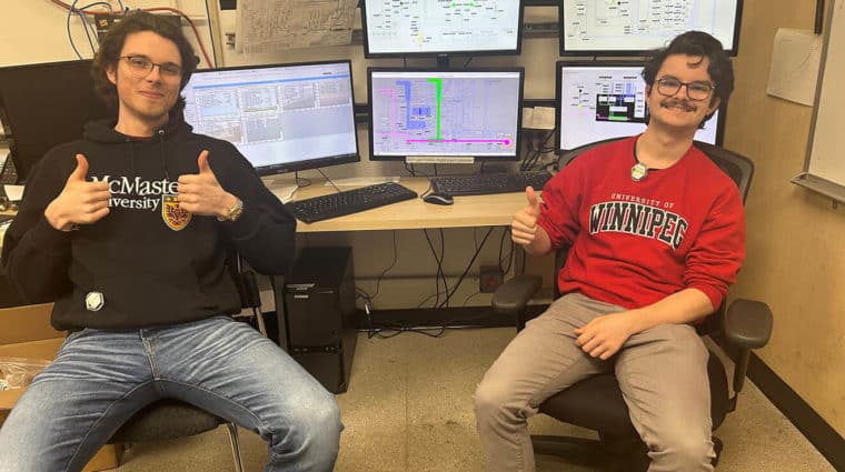 Colour photo of two people sitting in front of a desk with 6 control screens with circuit diagrams and alert flashes.