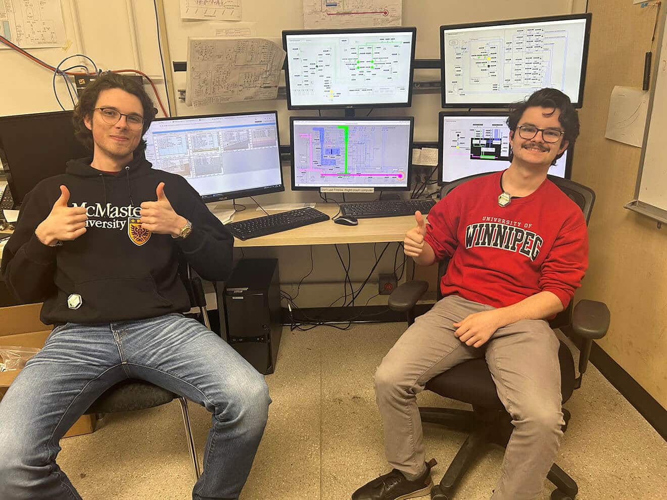 Colour photo of two people sitting in front of a desk with 6 control screens with circuit diagrams and alert flashes.