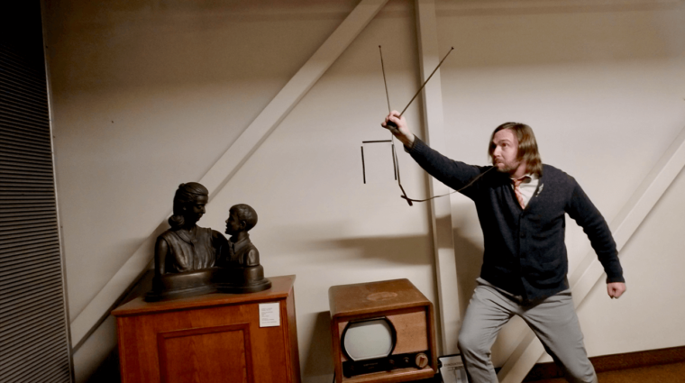 Long-haired, bearded man holding up a TV antenna with the wire in his mouth standing beside an old TV, and a bronze bust of a woman and young man.