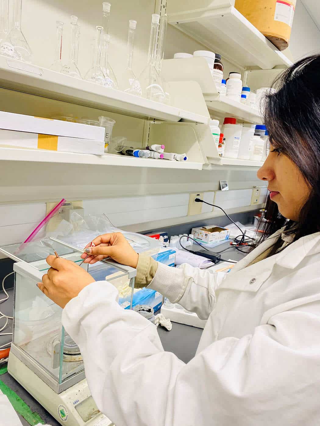 A researcher in a white lab coat working at a laboratory station, using tools to handle soil samples on a precision balance.