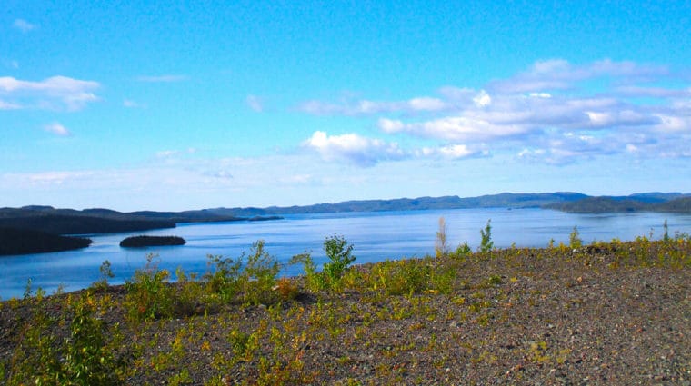 Photograph of a bright blue sky, bright blue lake, taken from a desolate brown hilltop, some trees on the opposite shoreline. It is a sunny day.