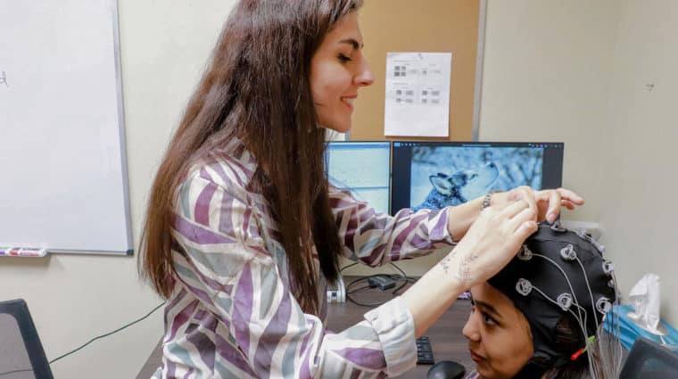 Researcher fitting an EEG cap onto a volunteer’s head as a husky image displays on a monitor, preparing for an emotion-focused study.
