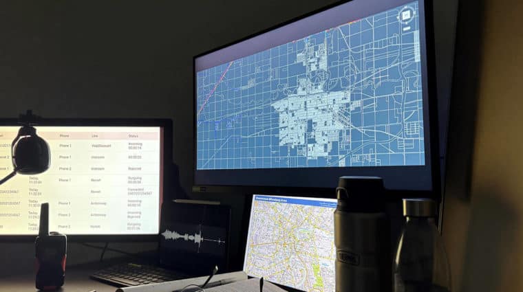 The image shows a dimly lit workspace with multiple screens displaying a city map, data logs, and an audio recording, tactical radio.
