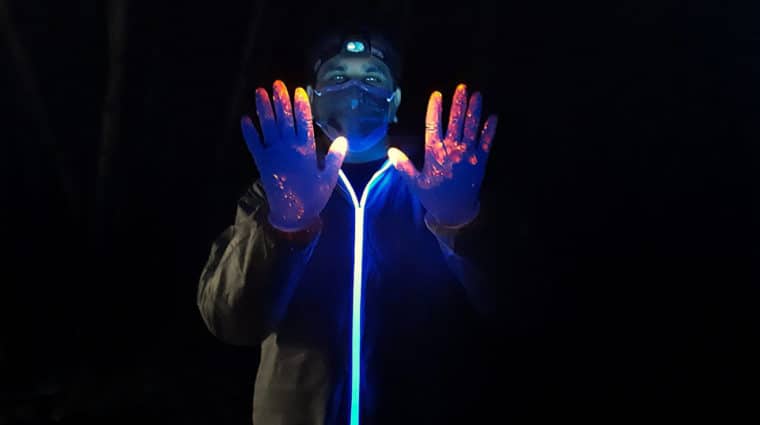 A person in a mask and plastic protective suit is covered in fluorescent powder, which is glowing under an ultraviolet light.