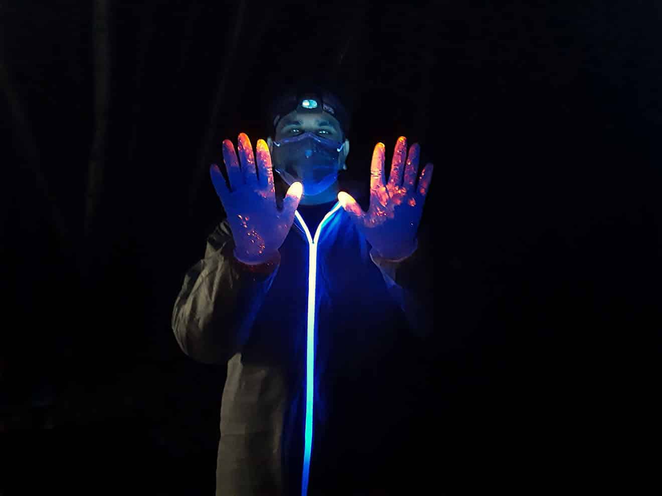 A person in a mask and plastic protective suit is covered in fluorescent powder, which is glowing under an ultraviolet light.