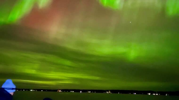 Researchers in a boat next to a yellow net in the water. The red and green northern lights are in the sky.
