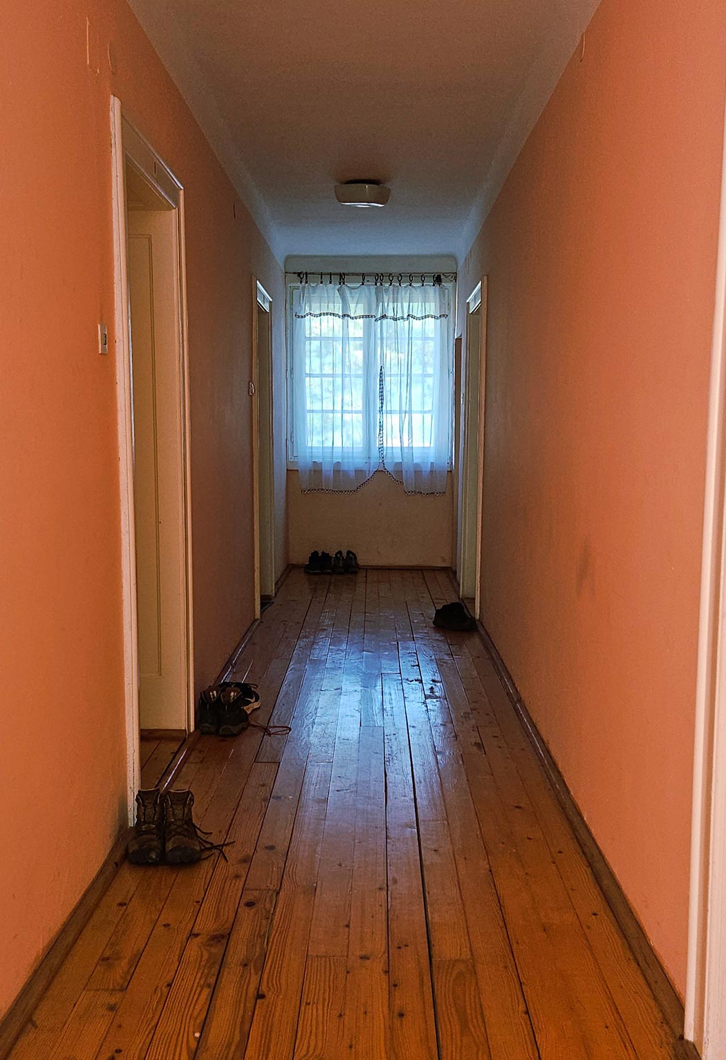 A hallway with pink walls, a bright window on the far wall and closed white doors each of which have work boots sitting on the hardwood floor.