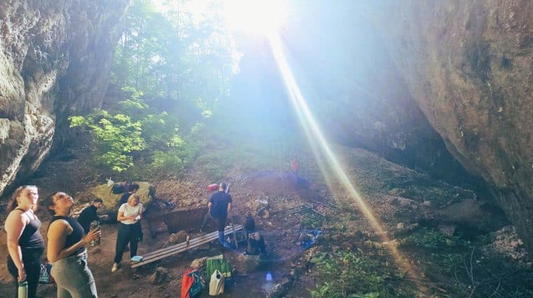 From the back of a rock shelter, an excavation site can be seen with team members setting up and the bright sun streaming rays onto the site