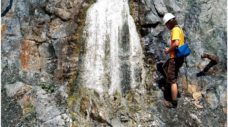 A researcher in orange standing on a rock wall in a mine collecting samples of a white mineral deposit flowing from the outcrop.